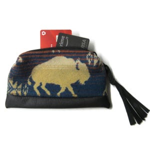 May include: A small, zippered coin purse with a black leather base and a patterned fabric top featuring a yellow bison silhouette against a blue and brown background. The purse has a black tassel and holds two credit cards.