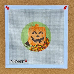 May include: A square needlepoint canvas with a light blue border and a stitched image of a jack-o'-lantern inside a green circle. The jack-o'-lantern is orange with a black face and a colorful garland. The text "PINPOINT" is at the bottom.