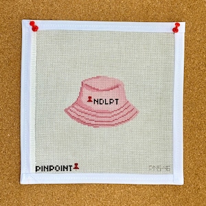 May include: A square needlepoint canvas with a pink bucket hat design and the text "NDLPT". The canvas is framed with white fabric and secured with red push pins. The text "PINPOINT" is at the bottom.