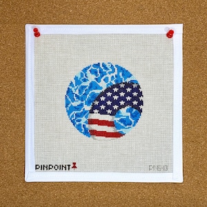 May include: A needlepoint canvas featuring a patriotic design. The artwork includes a blue circle with a pattern, and a section resembling the American flag with stars and stripes. The canvas is secured with red push pins.
