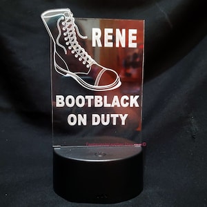 Könnte beinhalten: Eine transparente Acryl-LED-Lampe mit den weißen Worten "RENE BOOTBLACK ON DUTY". Eine detaillierte Umrisszeichnung eines Kampfstiefels befindet sich über dem Text. Die Lampe steht auf einem schwarzen Sockel.