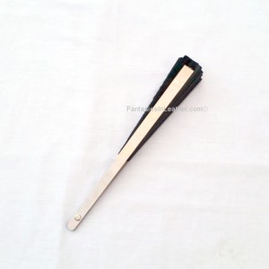 May include: A black and beige hand fan with a silver handle. The fan is folded and has a rectangular shape. The text "FantasiesInLeather.com" is visible on the fan.