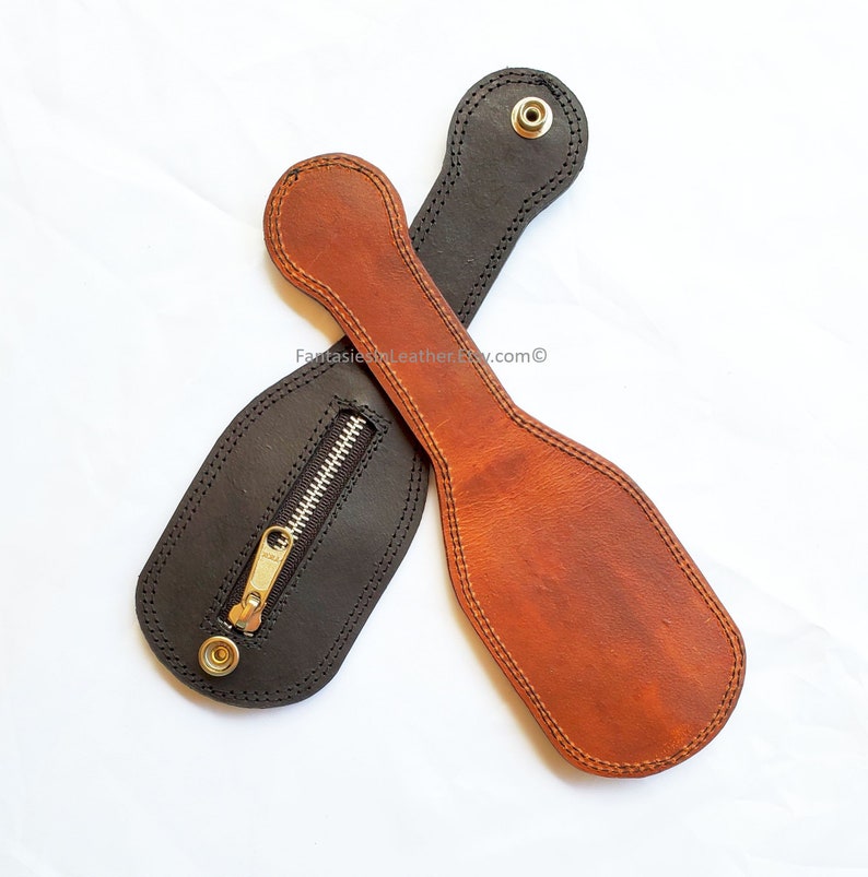 May include: Two leather paddles, one black and one brown, are displayed against a white background. The black paddle has a zippered pouch. Both paddles have a handle with a snap closure and are stitched around the edges.