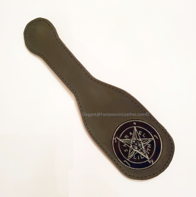 May include: A dark olive green leather paddle with a stitched handle and a circular emblem. The emblem features a pentagram and the text "SAMAEL LILITH". The product is from Elegant@FantasiesInLeather.com.