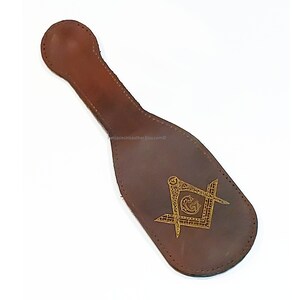 May include: A brown leather item with a handle and a decorative gold emblem featuring a square and compass design. The item has a stitched border and a rounded handle. The emblem is centered on the item.