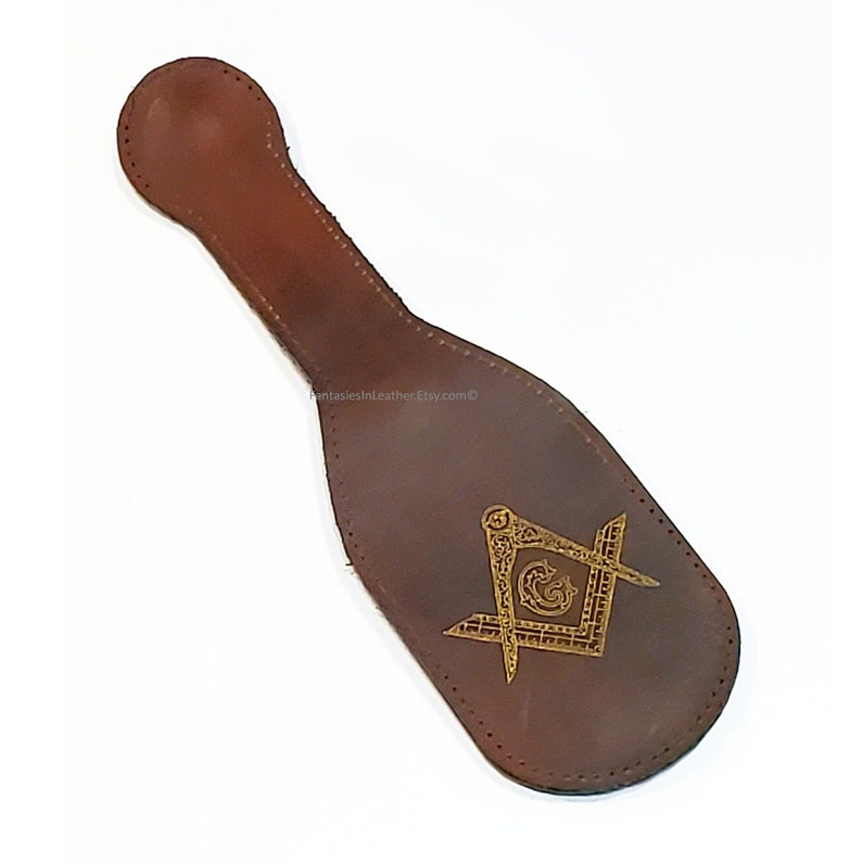 May include: A brown leather item with a handle and a decorative gold emblem featuring a square and compass design. The item has a stitched border and a rounded handle. The emblem is centered on the item.