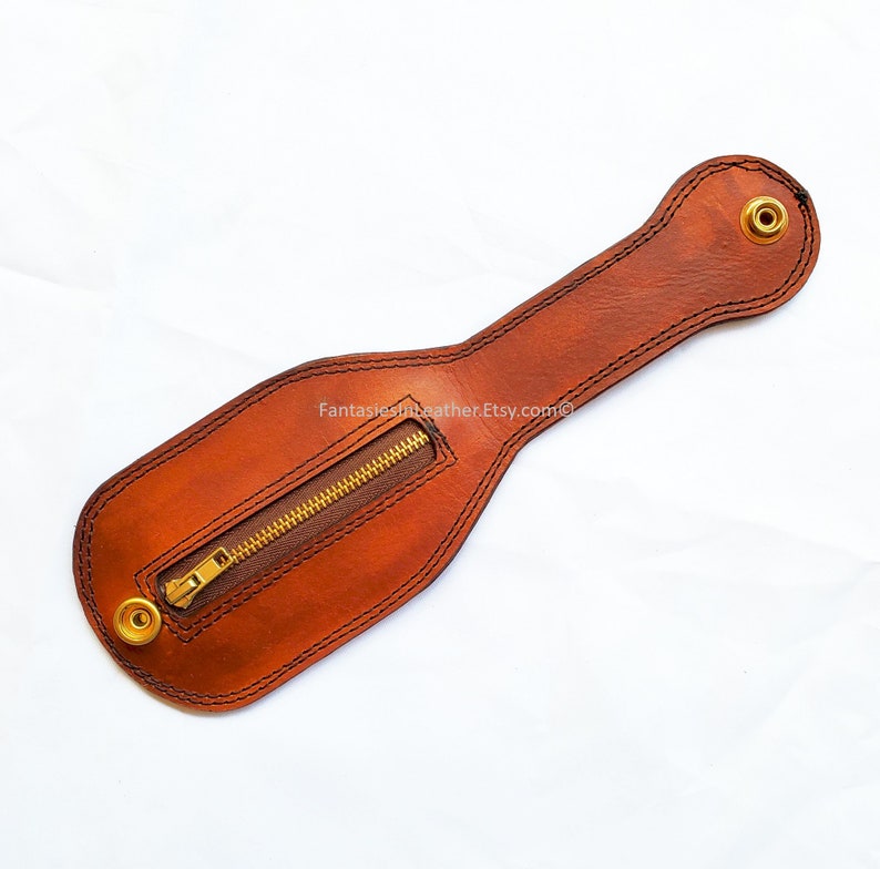 May include: A brown leather item with a zipper and brass rivets. The item has a long, narrow handle and a wider, rounded base. The zipper is gold-colored. The text "Fantasiesinleather.Etsy.com" is visible.