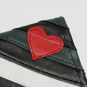 May include: Close-up of a small, triangular leather wallet. The wallet features a red heart applique on a black and green striped design. The wallet has a white stripe on the bottom.