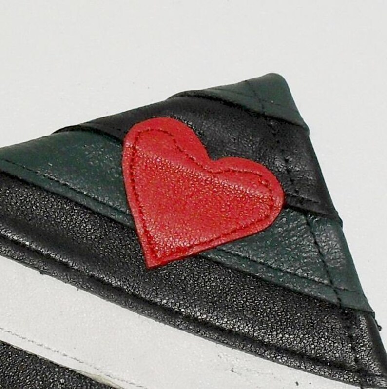 May include: Close-up of a small, triangular leather wallet. The wallet features a red heart applique on a black and green striped design. The wallet has a white stripe on the bottom.