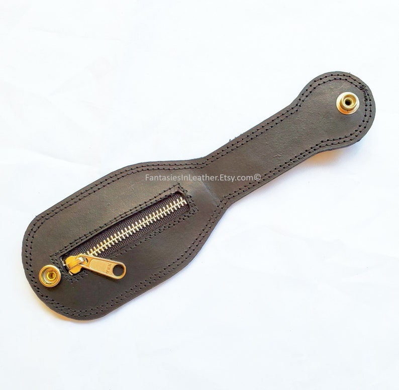 May include: Black leather pouch with a zipper closure and brass-colored rivets. The pouch has a stitched border and a long handle-like extension. The text "FantasiesInLeather.Etsy.com" is visible on the surface.