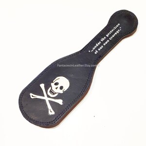 May include: Black leather paddle with a white skull and crossbones design. The paddle has a handle and the text "...under the protection of our own courage." is printed on the handle.