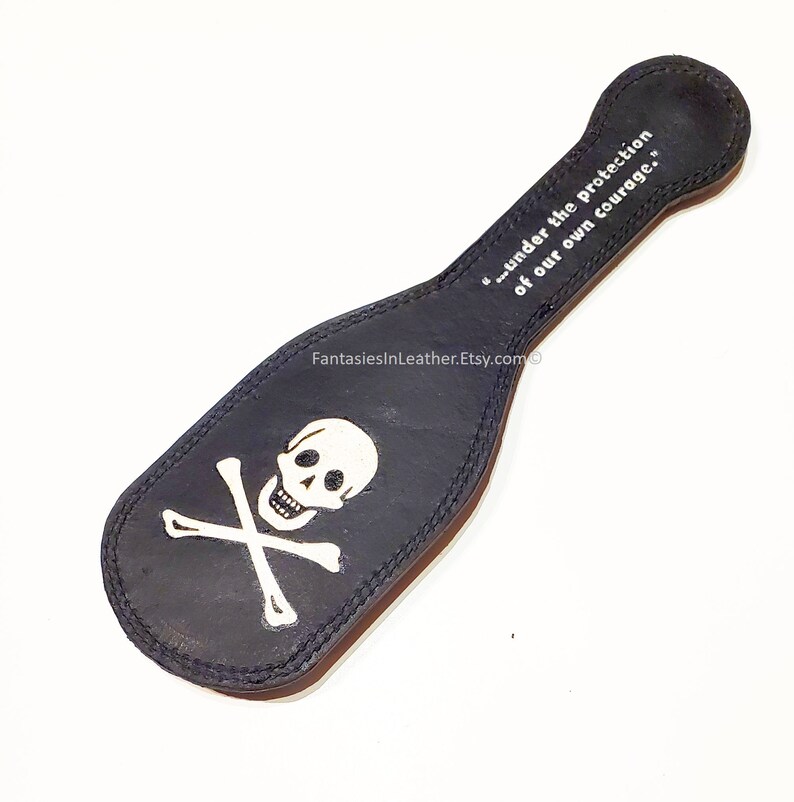 May include: Black leather paddle with a white skull and crossbones design. The paddle has a handle and the text "...under the protection of our own courage." is printed on the handle.