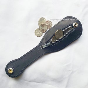 May include: A black leather coin purse with a zipper closure, holding several coins. The purse has a long, slender handle and a snap closure at the end. The text "Elegant@FantasiesInLeather.com" is visible in the image.