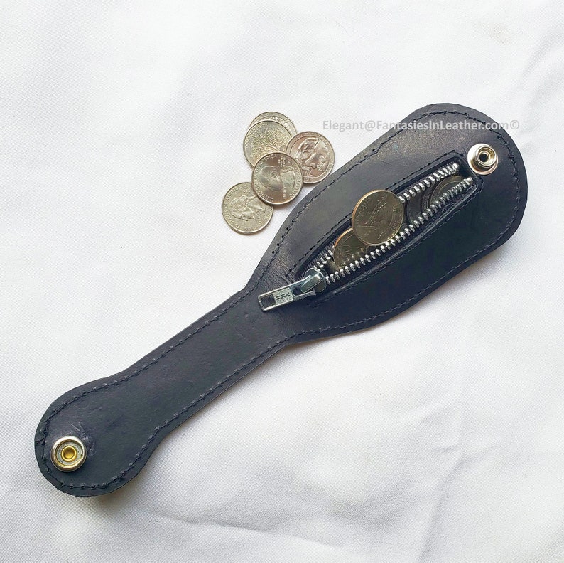 May include: A black leather coin purse with a zipper closure, holding several coins. The purse has a long, slender handle and a snap closure at the end. The text "Elegant@FantasiesInLeather.com" is visible in the image.