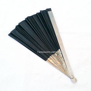 May include: A black hand fan with a silver metal handle and accents. The fan is made of pleated black material and is fanned out. The text "FantasiesInLeather" is printed on the fan.