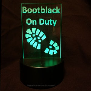 Könnte beinhalten: Ein rechteckiges, beleuchtetes Schild mit den Worten "Bootblack On Duty" in grün. Darunter zwei grüne Fußabdrücke. Das Schild steht auf einem schwarzen Sockel.