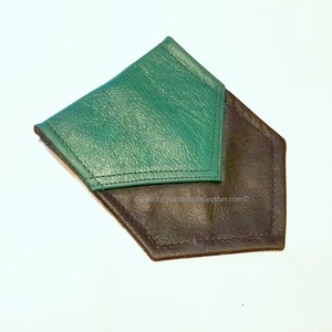 May include: A handmade leather wallet in a unique geometric shape. The wallet features a dark green flap over a black base, with visible stitching along the edges. The design is minimalist and modern, suitable for carrying cards and cash.