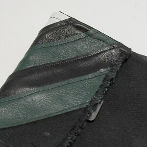 May include: Close-up of a black fabric item with a leather accent. The leather is black and dark green, with white accents. The item has a raw edge and a metal clasp.