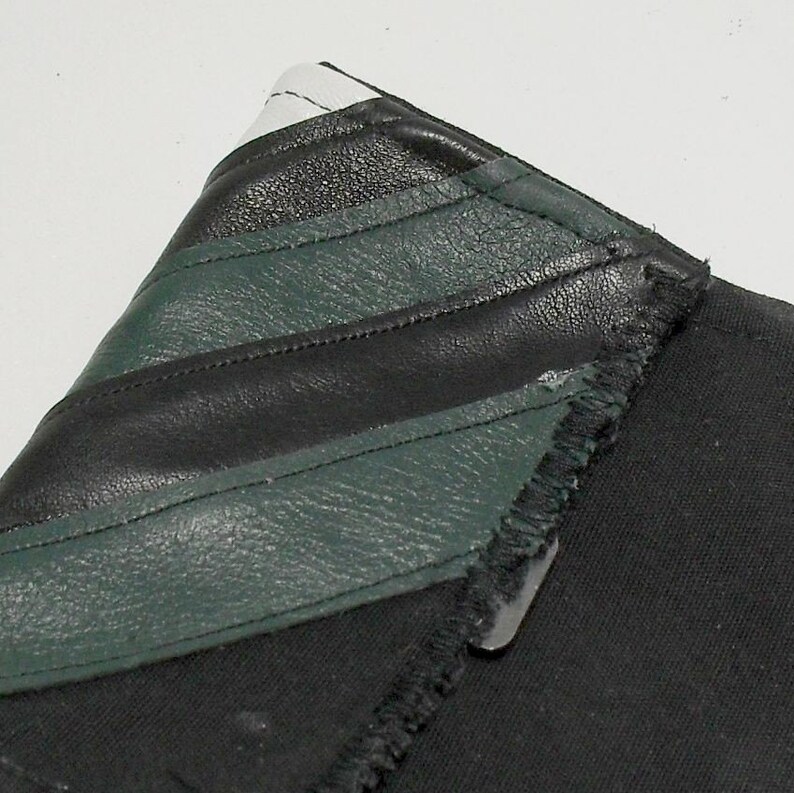 May include: Close-up of a black fabric item with a leather accent. The leather is black and dark green, with white accents. The item has a raw edge and a metal clasp.