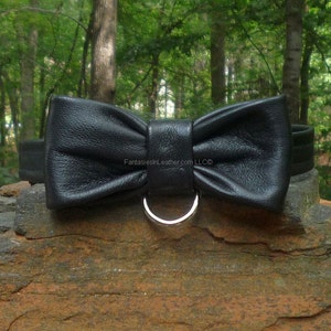 Könnte beinhalten: Eine schwarze Lederfliege mit einem silbernen Metallring. Die Fliege ist aus strukturiertem Leder gefertigt und hat eine klassische Bogenform. Der Text "FantasiesInLeather.com LLC©" ist sichtbar.