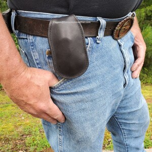 May include: A black leather belt pouch attached to a brown leather belt. The pouch is oval-shaped and appears to be made of smooth leather. The person is wearing blue jeans and a black shirt.