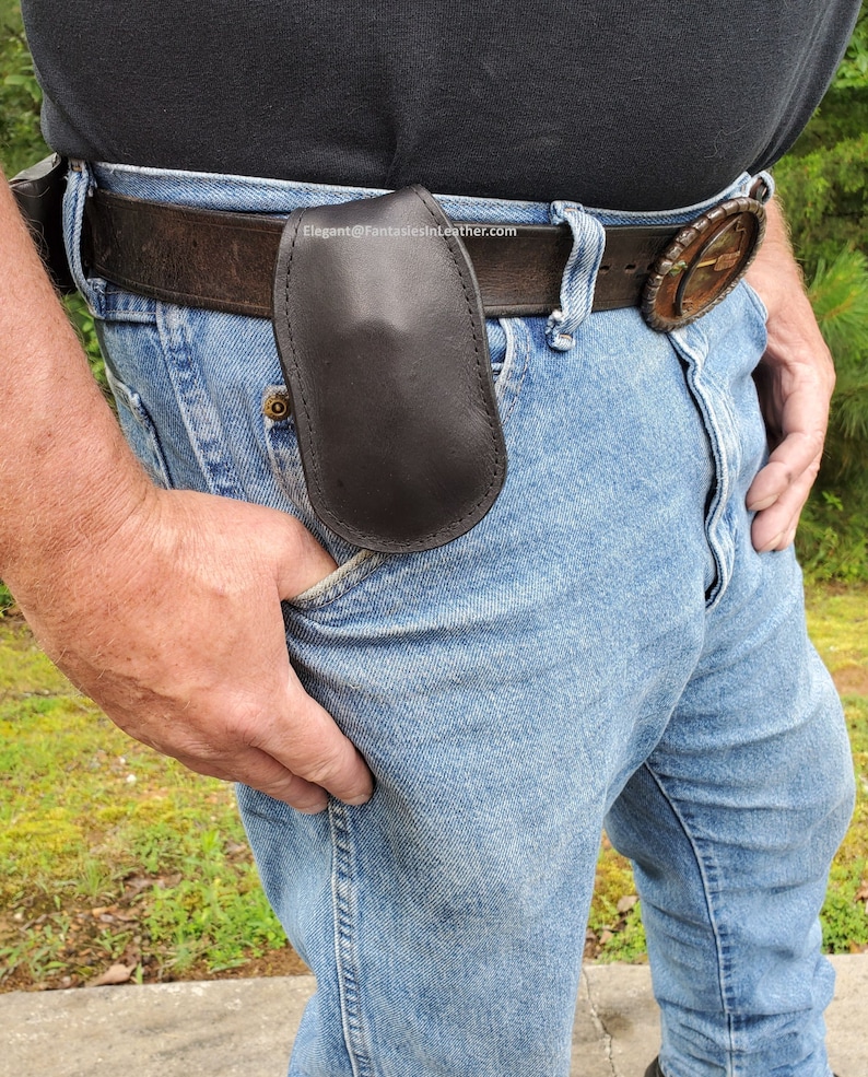 May include: A black leather belt pouch attached to a brown leather belt. The pouch is oval-shaped and appears to be made of smooth leather. The person is wearing blue jeans and a black shirt.