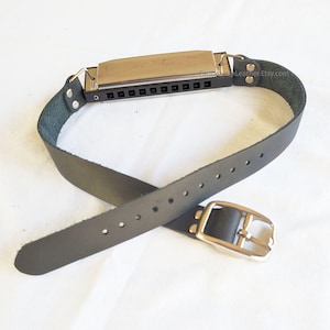 Pode incluir: Um cinto de couro preto com uma fivela prateada e uma gaita de boca presa. A gaita de boca é prateada com buracos pretos. O cinto é enrolado em volta da gaita de boca. O cinto tem vários furos para ajuste.