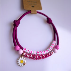 Puede incluir: Un brazalete de amistad morado con cuentas rosas y cuentas blancas con letras que deletrean "CHARLOTTE" y los números "015165081951". Un pequeño dije de margarita cuelga del brazalete.