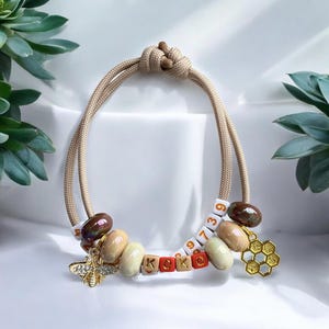 Può includere: Un braccialetto in corda beige con perline marroni, beige e rosse, un ciondolo a forma di ape dorata e un ciondolo a forma di favo dorato. Le perline hanno le lettere "KOKO" e i numeri "9739" su di esse.