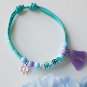 Peut inclure: Un bracelet ajustable bleu turquoise avec un charme en forme de fleur et un pompon. Le bracelet est composé de perles bleues, violettes et blanches avec le nom "WILMA" écrit en lettres blanches.