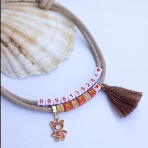 Puede incluir: Una pulsera de cordón beige con una borla marrón, un dije de oso dorado y dos filas de cuentas cuadradas. La fila superior muestra los números 0996138264 y un corazón, mientras que la fila inferior deletrea el nombre MARTINA con un corazón.