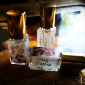 May include: Three small glass bottles with gold caps and labels. The bottles contain perfume oil. The labels have a vintage design with a picture of a woman and the text "Varda Psyche Perfume Oil".