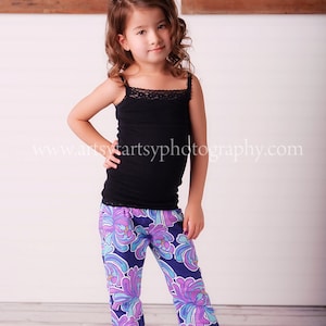 May include: A young girl wearing a black tank top with lace trim and floral print pants. The pants are primarily navy blue with a pattern of purple and turquoise flowers. The pants have a ruffled hem. The website address www.artsyartsyphotography.com is visible.