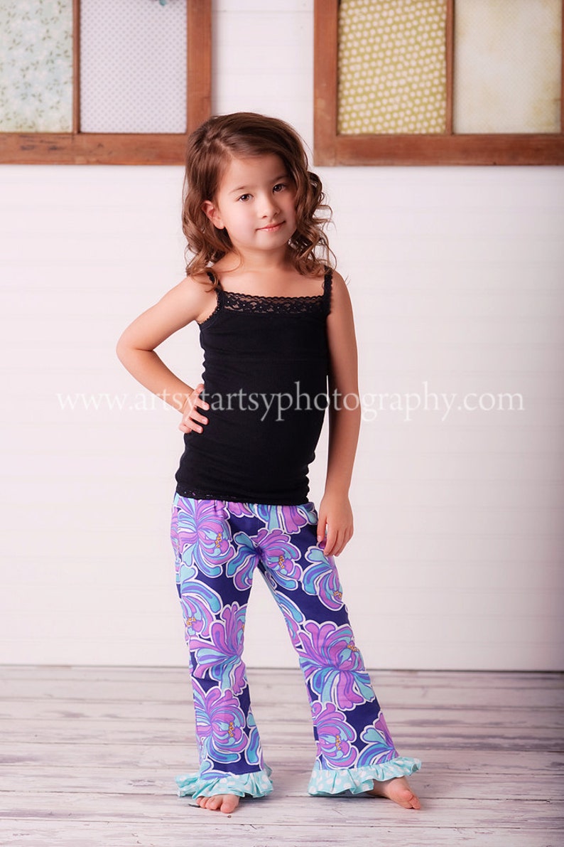 May include: A young girl wearing a black tank top with lace trim and floral print pants. The pants are primarily navy blue with a pattern of purple and turquoise flowers. The pants have a ruffled hem. The website address www.artsyartsyphotography.com is visible.