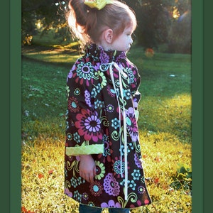 May include: A young girl wearing a brown and purple floral print dress with a green and white trim. She is wearing a yellow bow in her hair and is standing in a grassy field.