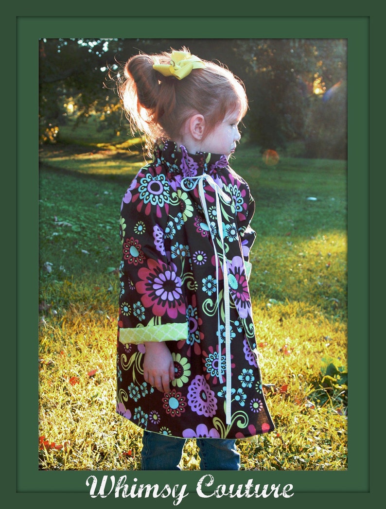 May include: A young girl wearing a brown and purple floral print dress with a green and white trim. She is wearing a yellow bow in her hair and is standing in a grassy field.