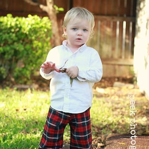 May include: A young child wearing a white button-down shirt and red and black plaid pants. The child is standing in a grassy area with a wooden fence in the background.