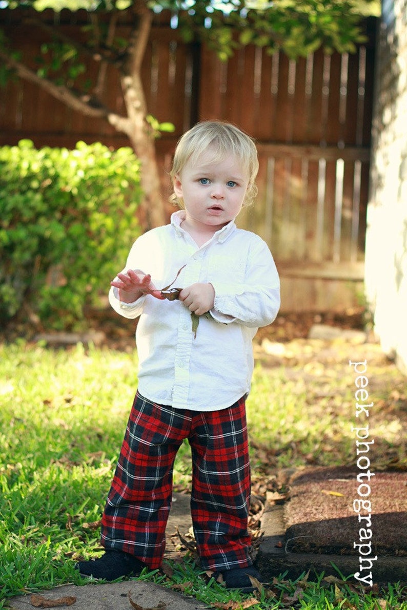 May include: A young child wearing a white button-down shirt and red and black plaid pants. The child is standing in a grassy area with a wooden fence in the background.