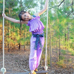 May include: A young person wearing a purple ruffled top and matching pants with a floral print is swinging on a wooden swing set in a wooded area.