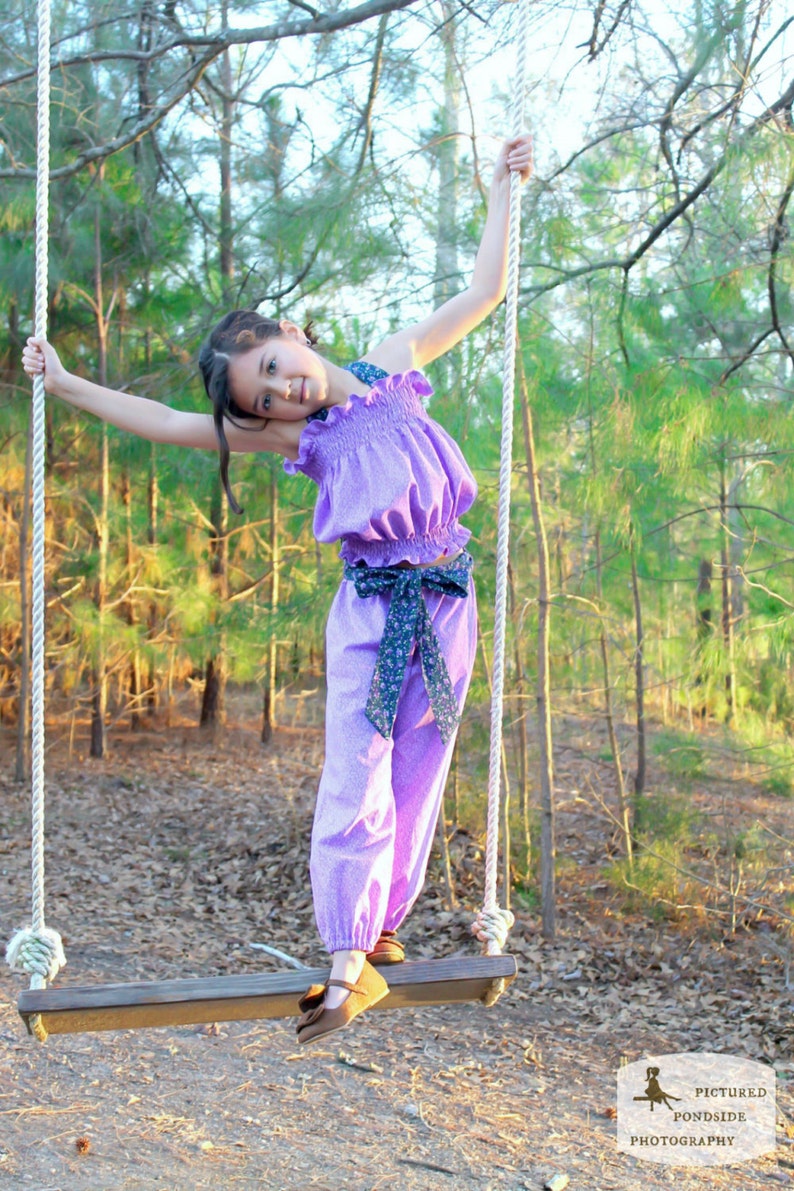 May include: A young person wearing a purple ruffled top and matching pants with a floral print is swinging on a wooden swing set in a wooded area.