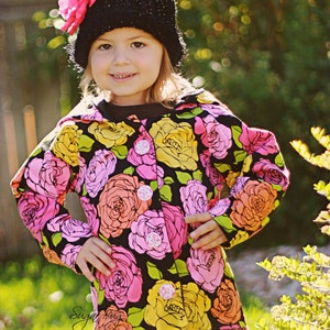 May include: A young girl wearing a black knit hat with a pink flower on top, a floral print jacket, and black and white polka dot pants. The jacket has a black background with pink, yellow, and green flowers.