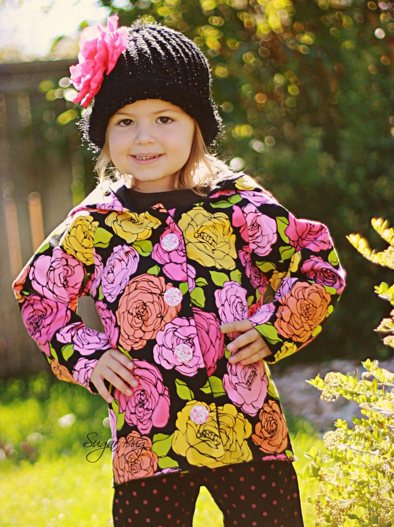 May include: A young girl wearing a black knit hat with a pink flower on top, a floral print jacket, and black and white polka dot pants. The jacket has a black background with pink, yellow, and green flowers.