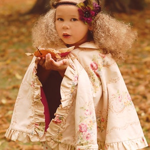 Op de afbeelding: Een jong meisje draagt een crèmekleurige bloemencape met ruches en een broek met stippen. Ze houdt een bruin blad in haar handen en heeft een bloemenband om haar hoofd.
