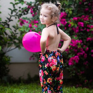 May include: A young girl wearing a black floral jumpsuit holds a pink balloon in front of a pink flowering bush.