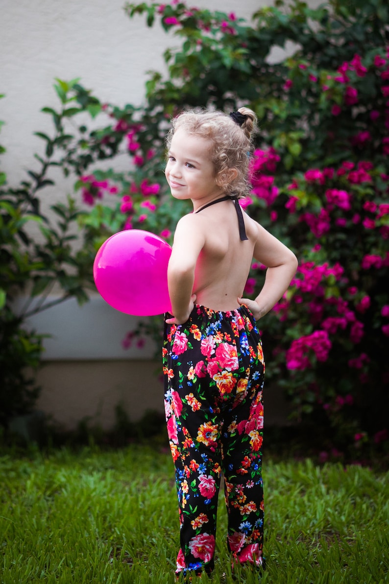 May include: A young girl wearing a black floral jumpsuit holds a pink balloon in front of a pink flowering bush.