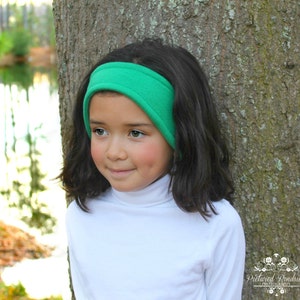 May include: A young person wearing a green fleece ear warmer headband. The headband is folded over the ears to keep them warm. The person is wearing a white turtleneck shirt.