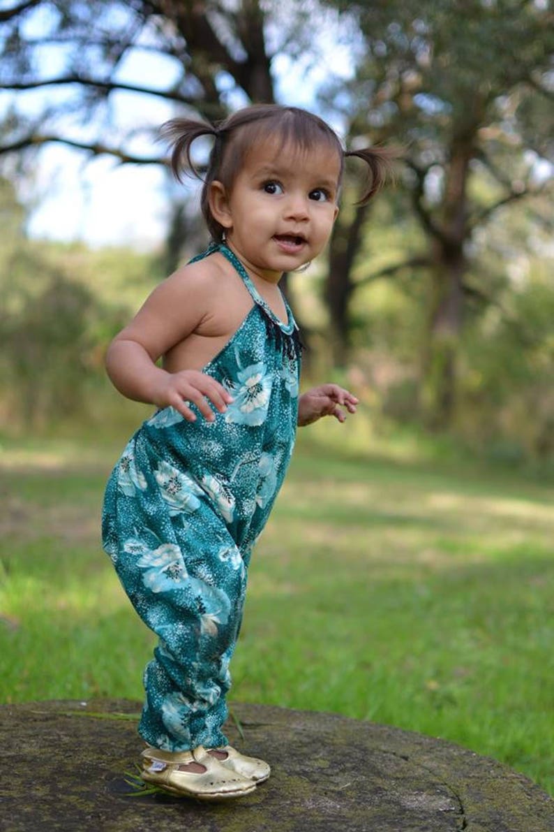 May include: A young girl with pigtails wears a teal floral jumpsuit and gold sandals. She stands on a stone surface in a grassy area.