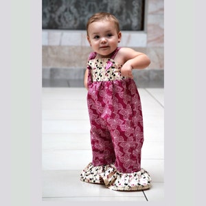 May include: A young child wearing a pink and white floral print romper with a ruffled hem. The romper has a small pink flower on the front.