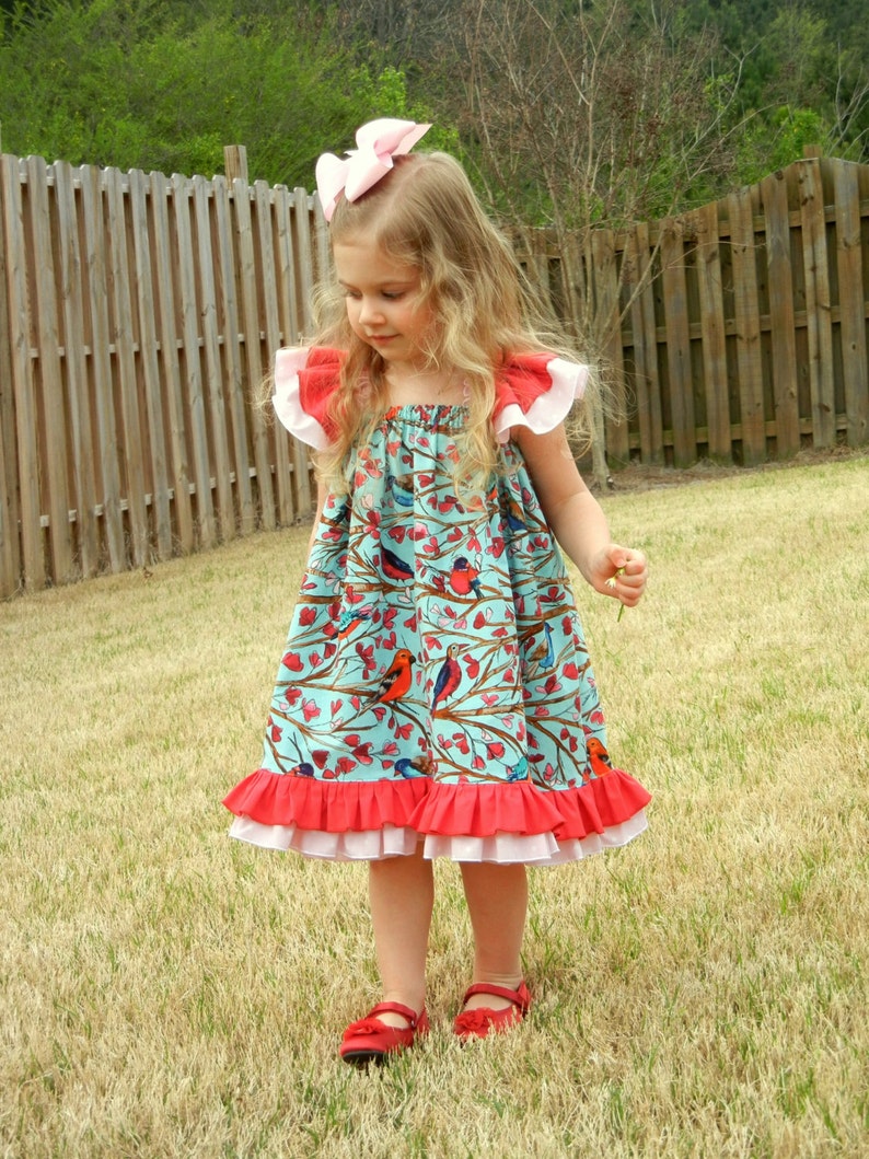 May include: A young girl wearing a turquoise dress with a bird and branch pattern. The dress has red and white ruffled trim and matching red shoes. A large pink bow adorns her hair.
