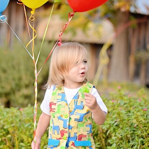 Könnte beinhalten: Ein kleines Kind trägt eine gelbe Weste mit einem bunten Tiermuster und blaue Jeans und hält einen Strauß Ballons fest. Die Ballons sind rot, gelb und blau.
